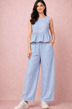 A woman modeling a sleeveless light blue and white gingham peplum top with matching wide-leg trousers, paired with white sneakers, against a solid dusty pink studio backdrop.
