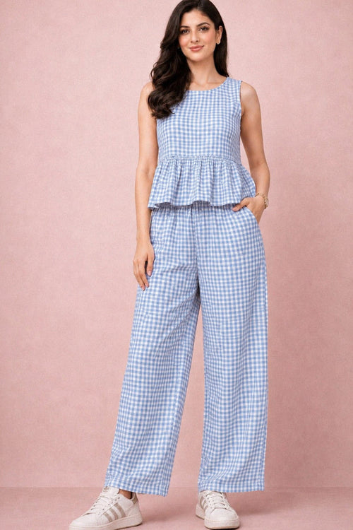 A woman modeling a sleeveless light blue and white gingham peplum top with matching wide-leg trousers, paired with white sneakers, against a solid dusty pink studio backdrop.